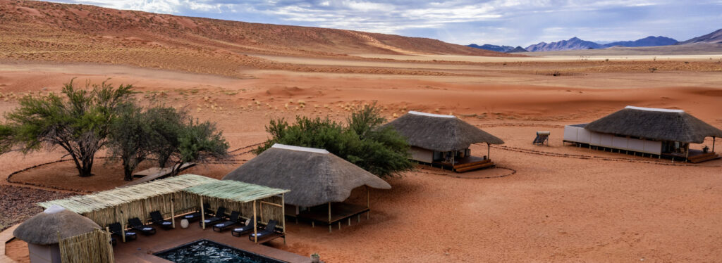 Aerial view of thatched lodge buildings and a pool deck set amid red desert dunes and distant mountains, Kwessi Dunes.