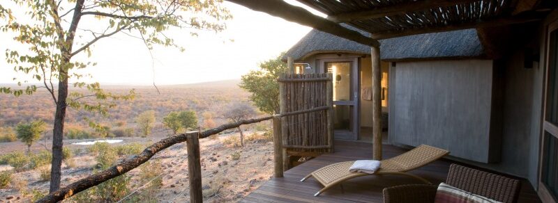 Thatched veranda with wicker furniture and an outdoor shower, overlooking open bushveld at dusk.