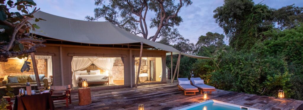 A canvas suite with a lit bedroom and freestanding bath, opening onto a wooden deck with a private lantern-lit plunge pool at dusk, Tembo Plains Zimbabwe.