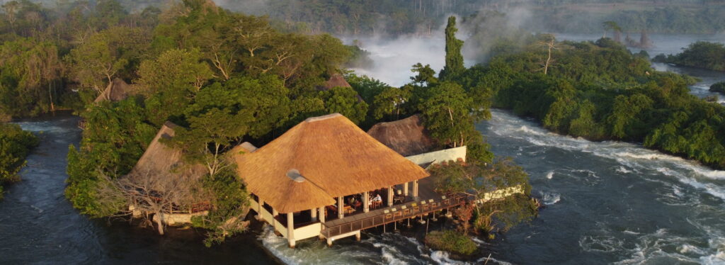 Aerial view of a thatched island lodge with a deck built over rushing rapids, surrounded by dense forest and misting waterfalls beyond.