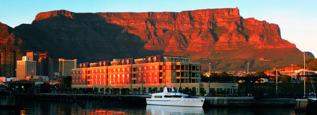 A waterfront hotel and moored yacht reflected in harbour water, with Table Mountain glowing red at sunset.