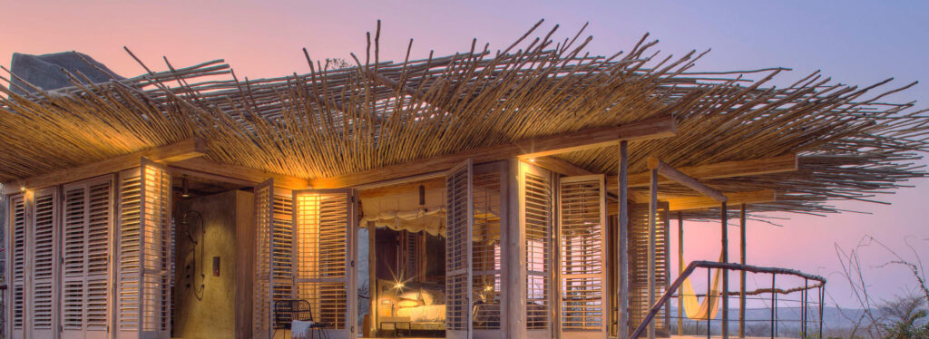 A suite with a twig roof and open wooden shutters revealing a lit bedroom, fronted by a sunken lounge deck with cushions at dusk.