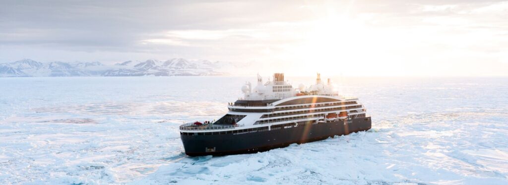 The expedition ship Le Commandant Charcot sits on calm water, reflected below, with snow covered peaks and glacial ice in the background.