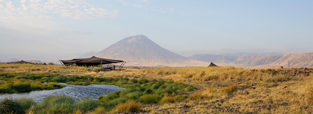 A canvas mess tent on open grassland beside a stream, with the volcanic cone of Ol Doinyo Lengai rising in the distance.