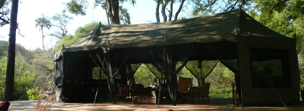 green canvas safari tent on a raised wooden deck, furnished with director's chairs and a writing desk, shaded by tall trees.