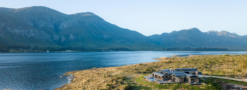 Aerial view of a curved roof lakeside house with a pool deck, set on a grassy shoreline beside a lagoon with mountains beyond.