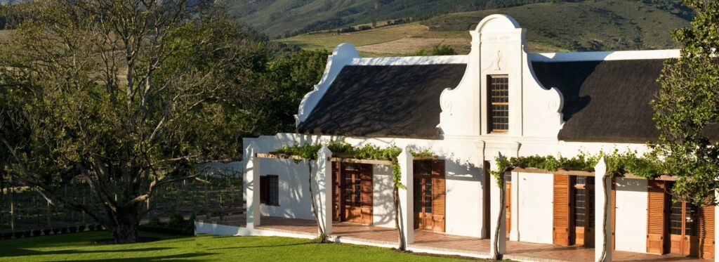 A white Cape Dutch manor house with a thatched roof, fronted by geese on a lawn with mountains behind.