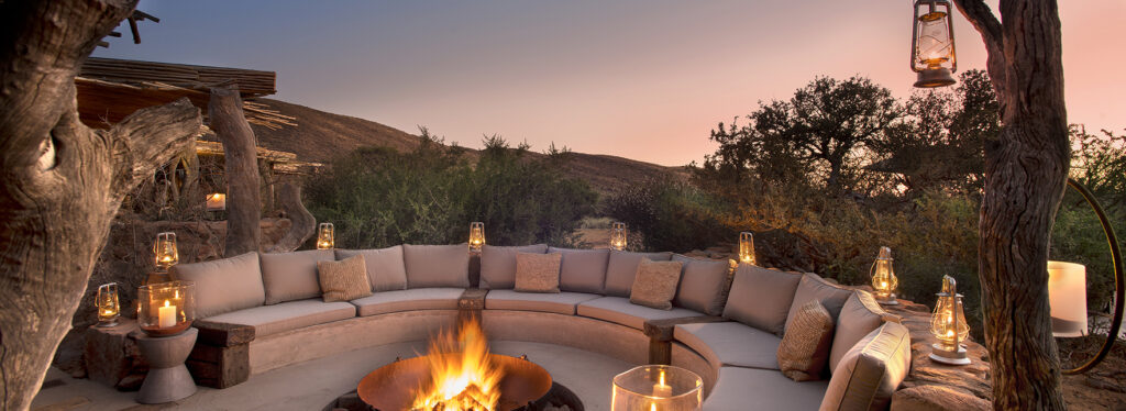 A curved lounge with cushioned seating around a fire pit, lit by lanterns at dusk.