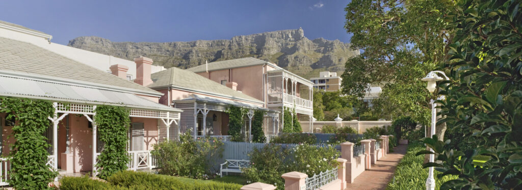 Pink Victorian style cottages with white lattice verandas along a brick pathway, bordered by hedges and a picket fence, with Table Mountain behind.