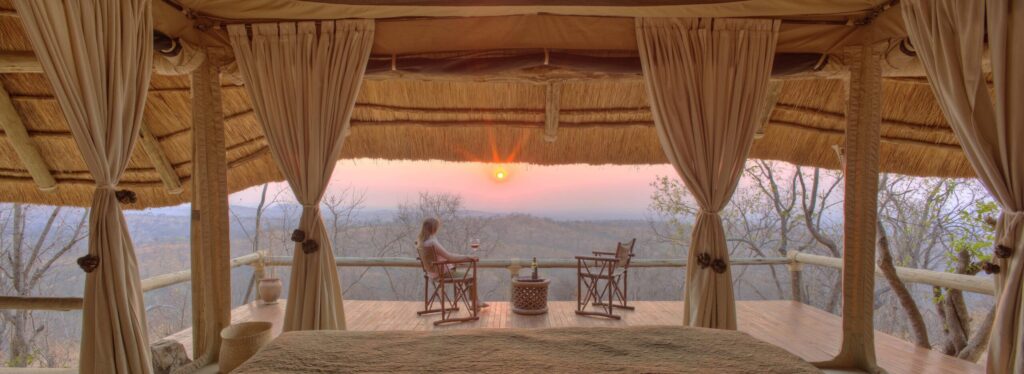 From a bed beneath a thatched roof, a guest sits on the deck watching the sunset over wooded hills, with a glass of wine on the table.