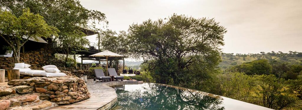 An infinity pool with a stone and timber deck, edged by loungers and umbrellas, overlooking wooded hills at sunrise, Faru Faru Lodge Tanzania.