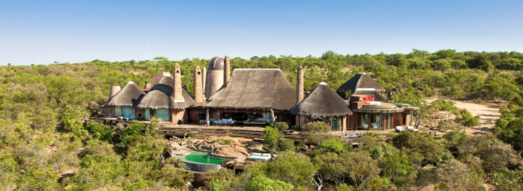 Aerial view of thatched, conical roofed lodge buildings with tall chimneys and an observatory dome, set among dense bushveld with a rock pool below.