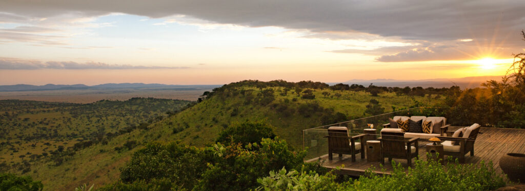 Singita Sasakwa Deck Overlooking the Serengeti. A wooden deck with lounge seating on a hilltop, overlooking rolling grassland stretching to the horizon under a cloudy sunset sky.