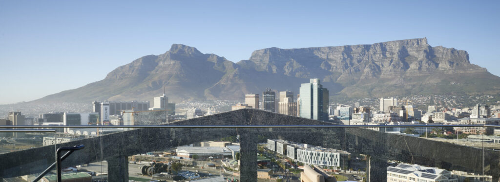 A rooftop terrace with lounge chairs and a glass balustrade, overlooking the Cape Town city skyline with Table Mountain rising behind.