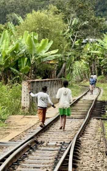 Children walking along a railway line through lush banana trees in Andasibe, with more people further down the tracks.