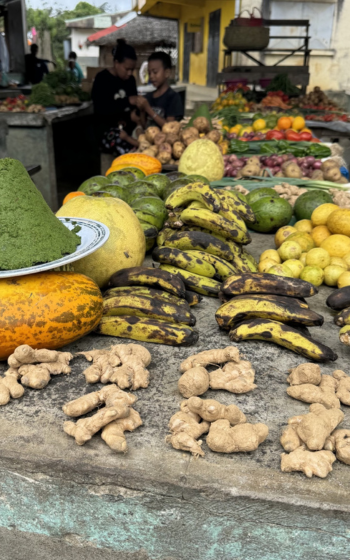 Roadside market stall piled with bananas, ginger, pumpkins, limes and avocados, with a mound of pounded green leaves on a plate.